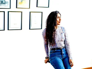A woman with dark long hair sits on a stool in a bright gallery space, wearing a patterned blouse and blue jeans, looking to the right. Framed artworks hang in two rows on the white wall behind her.