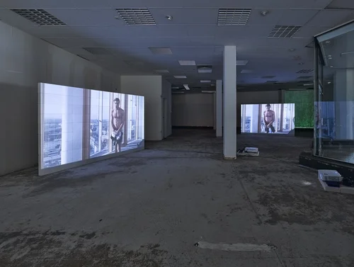 Film installations are shown on two large screens in an empty former factory space. The screens show a man holding items of clothing in front of his body.