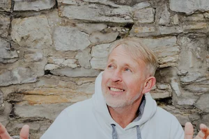 Das Bild zeigt einen Mann mit hellem Haar und Bart, der eine weiße Kapuzenjacke mit einem grafischen Design auf der Brust trägt. Es handelt sich um den Künstler Manfred Webel. Im Hintergrund sieht man den Dom zu Paderborn mit einer steinernen Fassade und einem grünen Dach.
