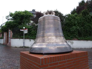 The picture shows a large, metal bell standing on a low brick pedestal. In the background are green trees and a building with a white wall and a gate.