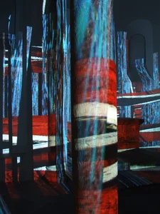 The picture shows a modern, abstract installation with luminous, colored light projections on cylindrical and rectangular structures. The scene is dark and dramatic, with light reflections that create a futuristic and dynamic ambience.