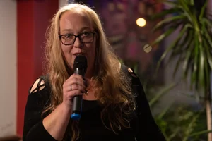 Julia Kluge, with long blonde hair and black glasses, is standing indoors and speaking into a microphone. She is wearing a black top with shoulder cut-outs and looks warmly into the camera; colorful lights and green plants can be seen in the background.