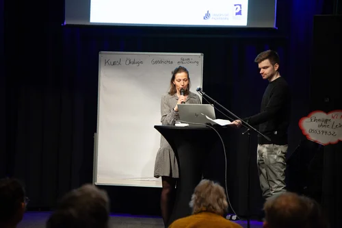 Auf dem Bild halten Brigitte Zacharias und Benedikt Heitmar auf der Bühne im Deelenhauseine Präsentation. Sie stehen vor einem Whiteboard mit den Wörtern "Kunst", "Ökologie", "Geschichte", und "Städtische Entwicklung".