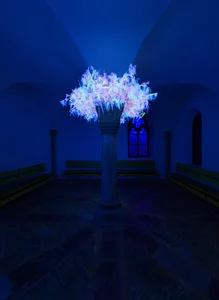 The picture shows a luminous, artistic installation that appears like a tree of light in the middle of the dark, blue-lit church. Pews and a colorfully illuminated window can be seen in the background, lending the scene a mystical atmosphere.