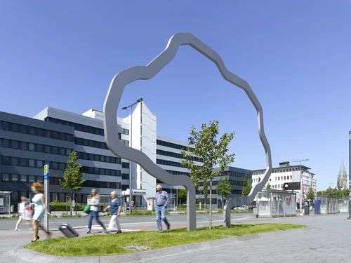 A tall metal sculpture, which forms the outline of a large tree, stands on a grass verge and surrounds a smaller tree. People walk past.