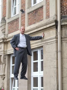 The picture shows a man in a suit who appears to be floating in front of the façade of a building. He is wearing a white shirt and a tie, and his posture suggests that he is leaning against the wall.