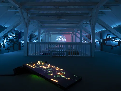 The image depicts a room with a wooden ceiling, illuminated by blue lights, featuring a lit rectangular object in the foreground and shadow figures on the walls.