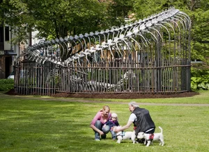 In a park there is a large sculpture showing a huge vertebral skeleton enclosed in a tall, curved metal cage. In the foreground, two adults are kneeling on a meadow with a small child and two small dogs.