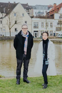 Ein Mann und eine Frau stehen nebeneinander vor einem kleinen Gewässer, hinter ihnen eine Reihe von Altbauten mit Fachwerkcharakter. Beide tragen helle Schals und Mäntel und blicken ruhig in die Kamera.