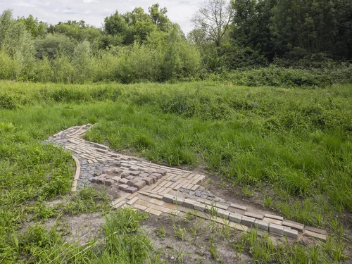 The picture shows an arrangement of paving stones reminiscent of a painting. The cobblestones are set into the ground and surrounded by tall grass.
