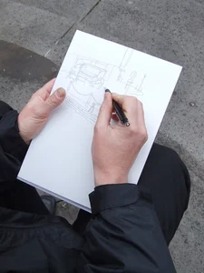 Photographed over the shoulder of a person, you can see hands drawing. The pencil sketch shows an urban scene.